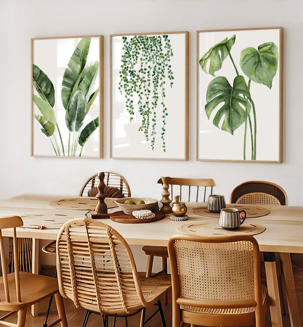 Three rectangular, light wood-framed prints featuring detailed watercolor illustrations of tropical plants are displayed above a long, light wood dining table. The prints depict, from left to right: a banana plant with large, textured leaves; a cascading hanging plant with small, round leaves; and a monstera plant with distinctive split leaves. The dining table is set with round woven placemats, dishes, and mugs, ready for a meal.