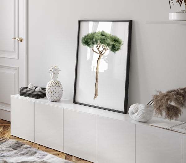 Mediterranean art is showcased with a framed watercolor illustration of an Italian Stone Pine leaning against a white wall on a white cabinet. The tree is depicted with green foliage and a brown trunk, framed in a thin black frame. On the cabinet, there is a black woven basket with two white spherical objects, a white pineapple-shaped vase, a white textured vase holding dried grasses, and a notebook. A portion of a wooden floor and a gray and white patterned rug are visible at the bottom.