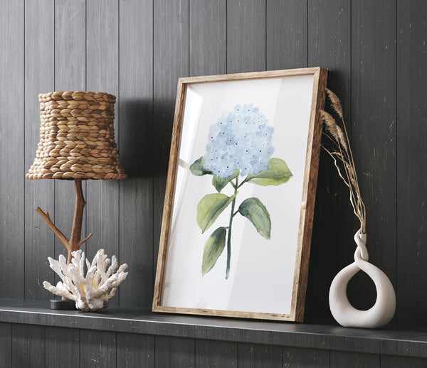 A framed watercolor painting of a blue hydrangea flower on a dark wooden shelf. A woven lampshade, a branch lamp, a coral decoration, and a white vase with a plant add visual interest to the scene.