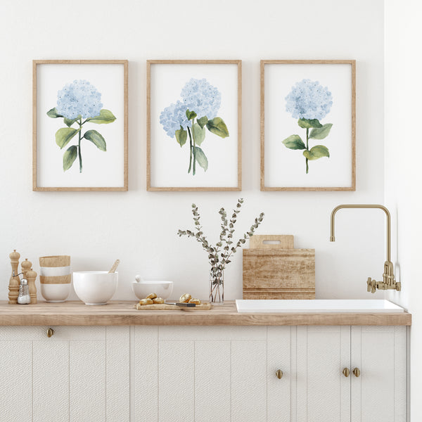 Three framed watercolor paintings of blue hydrangea flowers with green leaves and stems adorn a white wall above a kitchen counter. The counter is topped with a wooden cutting board, a bowl, and a vase with eucalyptus sprigs.