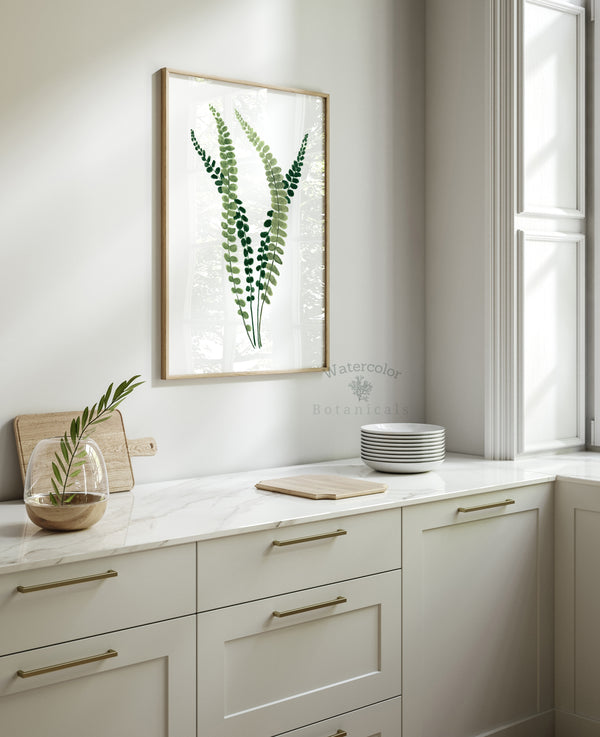 A framed watercolor painting showcases the intricate details of a fern, its small, soft green leaves rendered in subtle hues, creating a calming and elegant atmosphere above a kitchen counter with various kitchenware and fresh lemons.