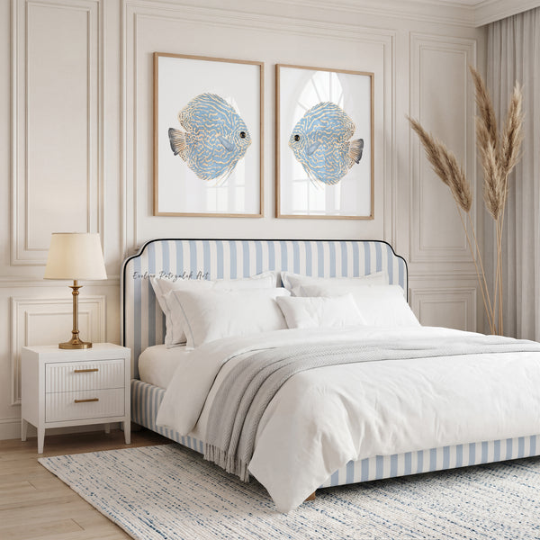 A styled bedroom scene featuring a bed with a light blue and white striped upholstered headboard and matching striped footboard. The room has white paneled walls, two framed blue fish artworks above the bed, a white nightstand with a lamp, and a tall pampas grass decoration near the window.