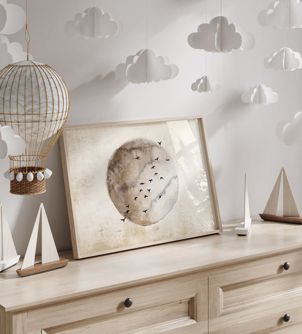 A framed watercolor painting of a moon in warm brown and beige tones with birds flying around it, hanging on a wall decorated with paper clouds and sailboats.