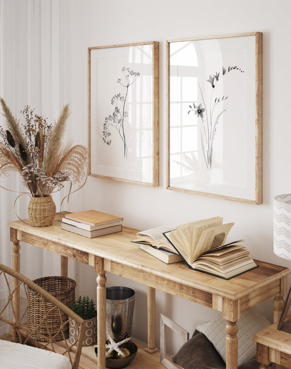 Two vertical framed botanical art prints with black ink sketches on white backgrounds, hanging on a light wall above a wooden console table styled with books and dried grasses.