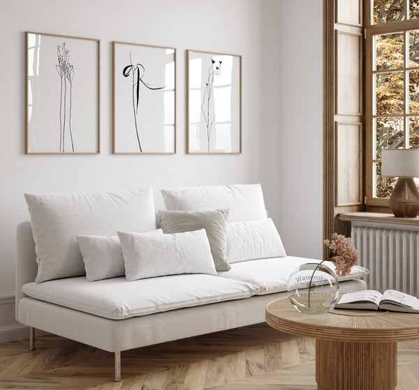 A set of three framed line drawings of various plants, rendered in sleek black lines against a crisp white background. These elegant botanical designs are displayed on a white wall above a comfortable-looking sofa in a living room setting. The overall aesthetic is minimalist and modern, with the artwork adding a touch of sophistication and tranquility to the space.