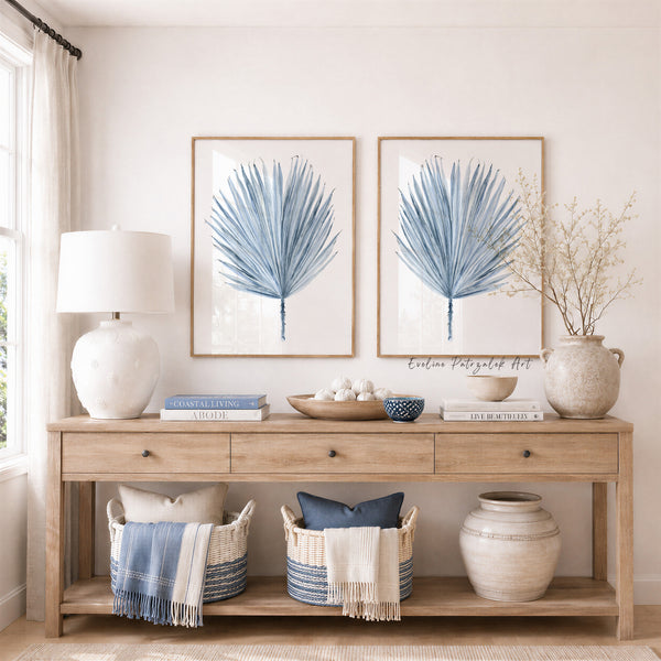 A styled interior scene showing two framed blue botanical prints with blue palm fronds hanging above a light wood console table. The room includes a white ceramic lamp, books, bowls, woven baskets with throw blankets, and ceramic vases, with a window and curtains visible on the left.