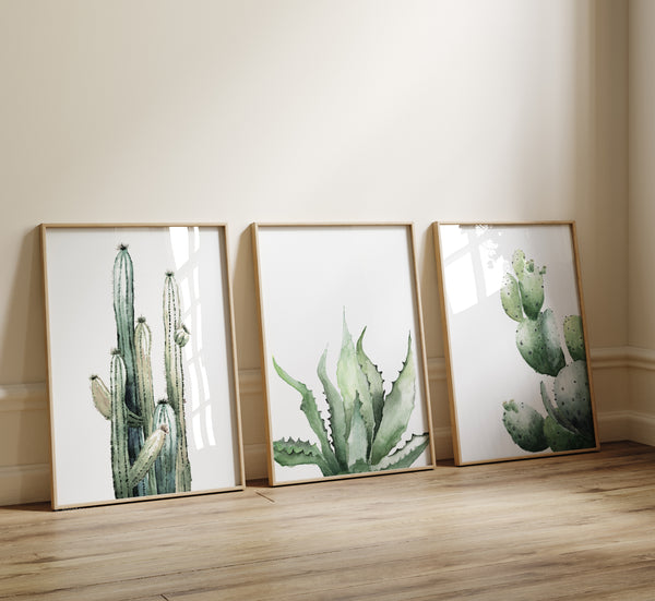 set of 3 watercolor prints featuring separately minimalist cactus opuntia, aloe vera and another tall cactus painted on white background stand in wooden frames leaning towards the wall