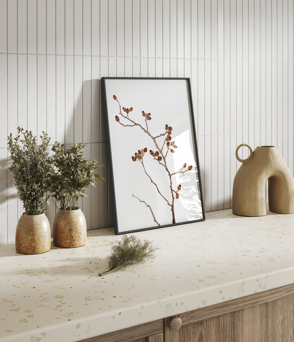 Single brown berry branch print in a black frame leaning on a kitchen counter