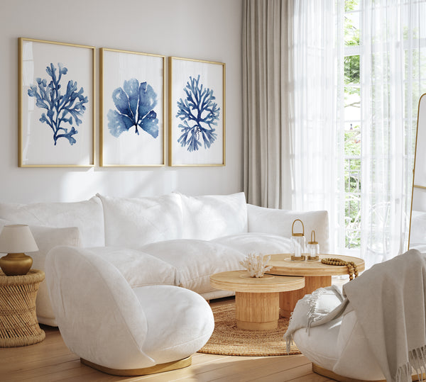 Watercolor paintings of minimalist seaweed and coral designs in deep navy blue add a touch of nature to a living room with a white sectional sofa, a round coffee table, and a white armchair.