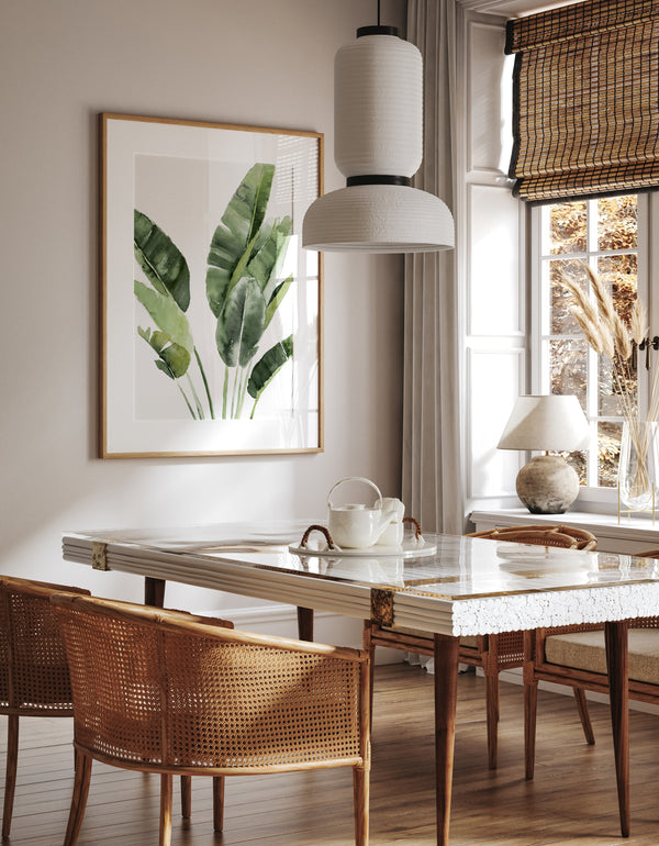 A watercolor painting of large green tropical banana leaves adds a touch of nature to a dining room with a long table, wicker chairs, and a hanging light fixture.