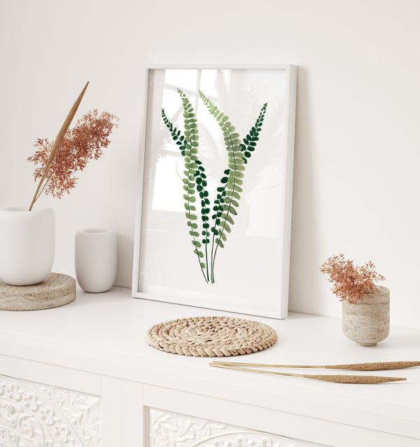 A framed watercolor painting showcases the intricate details of a fern, its small, soft green leaves rendered in subtle hues, complementing a space with a white bench, a vase of dried pampas grass, and decorative accents.