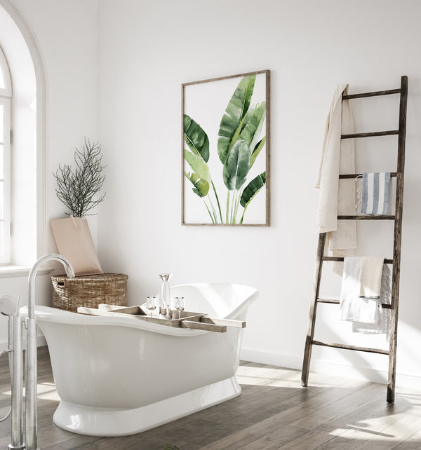 A watercolor painting of large green tropical banana leaves adds a touch of nature to a bathroom with a bathtub, a wooden ladder, and a white wall.