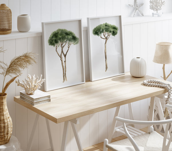 Mediterranean art is showcased in a workspace with two framed watercolor illustrations of Italian Stone Pines on a light-colored wooden desk against a white shiplap wall. The trees are depicted with green foliage and brown trunks, each in a thin white frame. On the desk, there is a white vase, a string of white beads, and a small stack of books with a piece of coral on top. To the left, a woven vase holding dried grasses sits on the floor. To the right, a white lamp with a beige shade is visible.