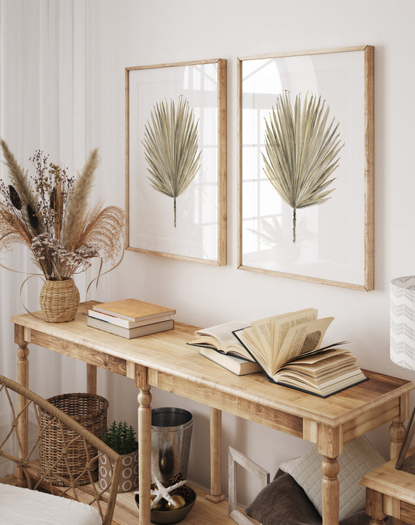 Two large-scale botanical prints of palmetto leaves, each framed in a natural wood frame, hang on a white wall above a wooden console table. The table is styled with a vase of dried flowers, a stack of books, and decorative objects, creating a cozy and inviting atmosphere. 