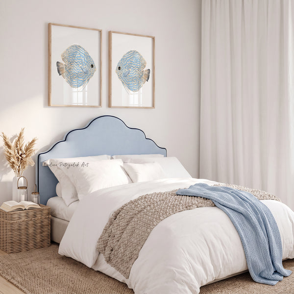 A styled bedroom scene featuring a light blue upholstered bed headboard with a curved scalloped top, dressed in white bedding, a beige knit throw, and a light blue blanket. Two framed fish wall art prints hang above the bed against a white wall.