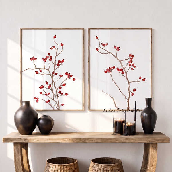 Two framed botanical wall art prints with red berries on branches, displayed above a wooden console table with dark vases and candles in a bright interior setting.