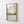 A framed art print featuring an abstract landscape with soft green and white clouds over a calm water scene, displayed on a white shiplap wall. The print is housed in a light natural wood frame, and a watermark reading 'Eveline Pietrzalek Art' is visible at the bottom.