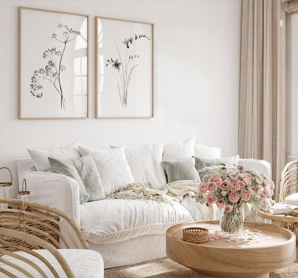 A bright, modern living room featuring a white textured fabric sofa with light sage green pillows, a round wooden coffee table with a pink rose bouquet, and two minimalist black and white botanical prints in light wood frames on the wall.