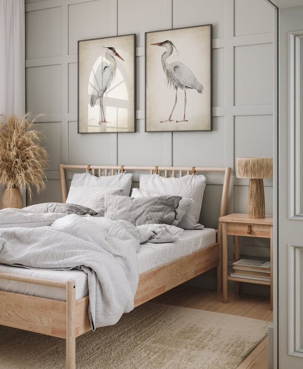 A pair of framed watercolor paintings depicting elegant great blue herons in soft neutral tones against a vintage-inspired background. The paintings are displayed on a wall above a bed in a bedroom with a serene and calming atmosphere.