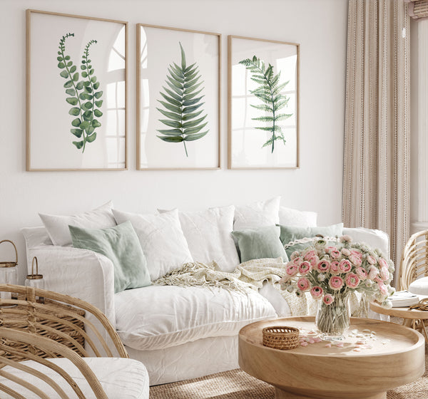 Three framed botanical prints with green leaf illustrations hang on a white wall above a white sofa styled with green and white pillows, a beige throw blanket, and a pink rose arrangement on a round wooden coffee table.