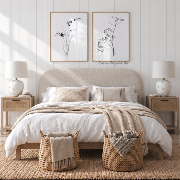 A styled bedroom scene featuring a bed dressed in white bedding and a beige throw blanket, with two matching bedside tables and white lamps on either side. Above the bed are two framed botanical prints on a white shiplap wall.