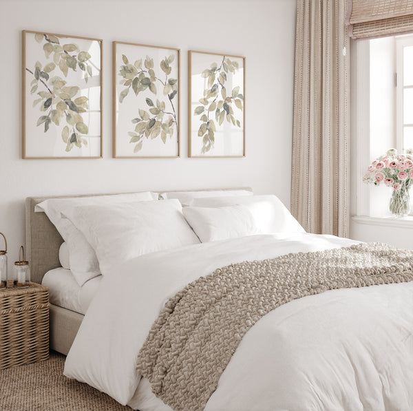 A triptych of watercolor paintings featuring delicate branches and leaves in soft shades of green and brown. The paintings are framed and displayed on a white wall above a bed.  The artwork creates a calming and elegant atmosphere in the bedroom.