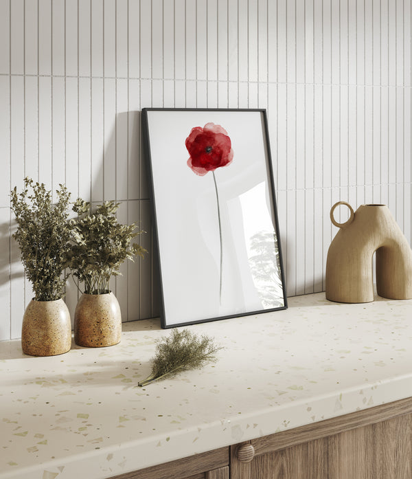 A framed watercolor painting of a single red poppy flower, displayed on a countertop with a textured surface. The painting is framed in a simple black frame, adding a touch of contrast to the overall aesthetic. The presence of small potted plants and decorative vases creates a warm and inviting atmosphere.