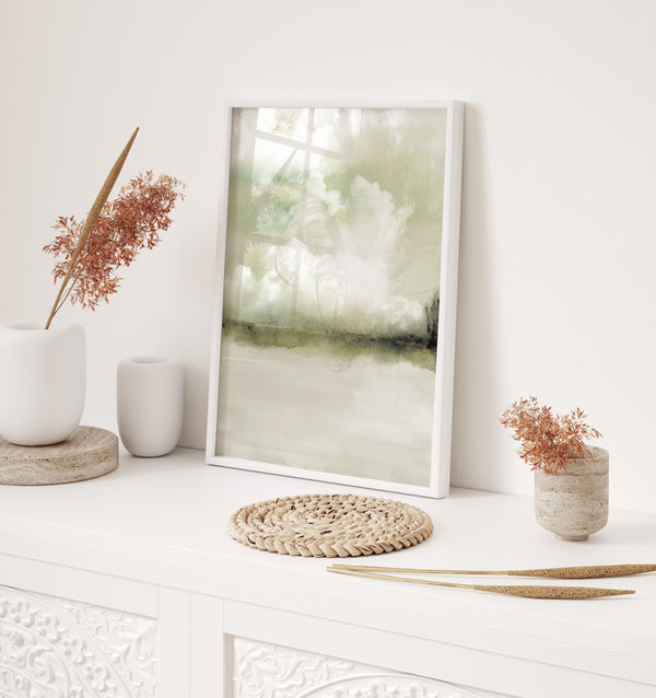 A framed abstract landscape art print with soft green and beige tones, styled on a white console table with dried plants and neutral home decor accessories.