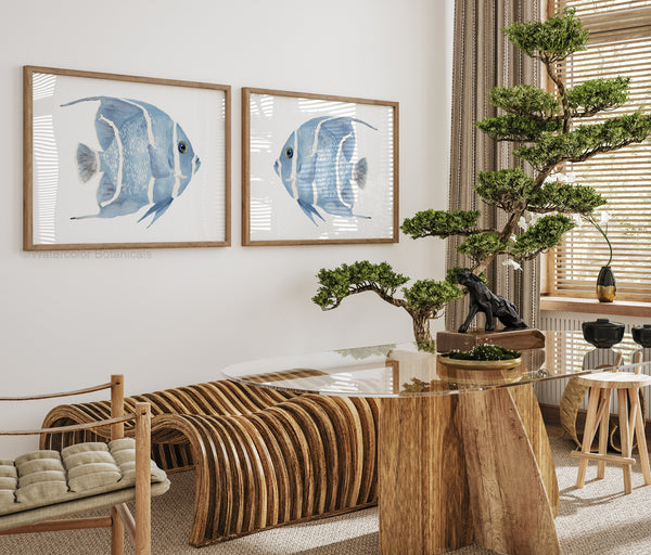 Two watercolor paintings of blue angelfish, each framed in a natural wood frame. The fish are facing each other in a horizontal orientation, creating a sense of balance and symmetry. The paintings are displayed on a white wall above a dining table with wooden chairs and a glass tabletop. The room is decorated with natural elements like woven baskets, dried flowers, and a small potted plant, creating a warm and inviting atmosphere.
