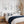 A styled bedroom scene with a bed dressed in white bedding and a navy knit throw, flanked by two wooden nightstands with white lamps. Above the bed are three framed botanical prints with blue rosehip branches 