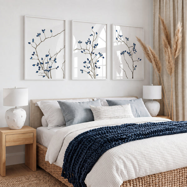 A styled bedroom scene with a bed dressed in white bedding and a navy knit throw, flanked by two wooden nightstands with white lamps. Above the bed are three framed botanical prints with blue rosehip branches 
