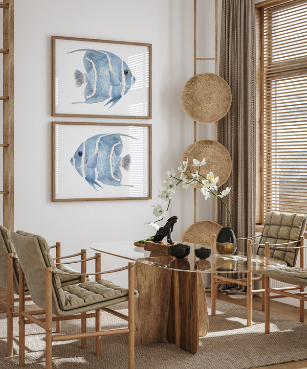 Two watercolor paintings of blue angelfish, each framed in a natural wood frame. The fish are facing each other in a horizontal orientation, creating a sense of balance and symmetry. The paintings are displayed on a white wall in a living room with dried flowers and decorative vases adding a touch of natural beauty to the space. The overall setting suggests a coastal or beach-inspired theme.