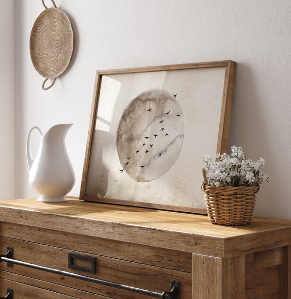 A framed watercolor painting of a moon in warm brown and beige tones with birds flying around it, hanging on a wall above a wooden dresser with a pitcher and a basket of flowers.