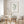 A watercolor painting of green sage leaves adds a touch of nature to a dining room with a round table, white chairs, and a white marble vase with dried branches.