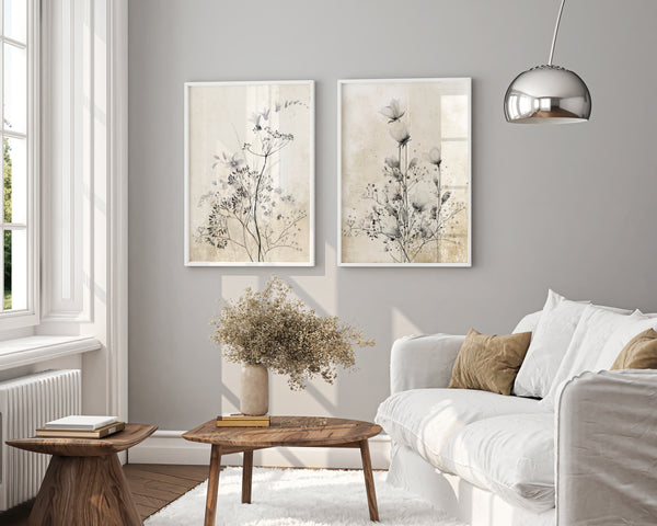 A pair of framed botanical prints featuring delicate black roses and other flowers in shades of grey and white hang above a cozy sofa in a living room with a neutral color palette.