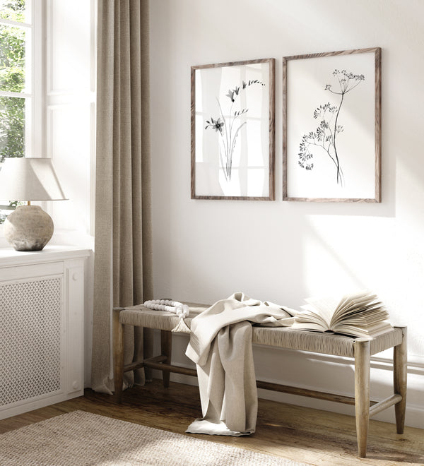 A styled interior scene with a wooden bench draped in beige fabric and an open book, beneath two framed botanical prints on a white wall. A window, beige curtain, table lamp, and rug are visible in a bright room.