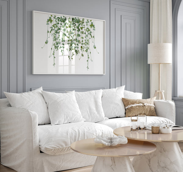 A watercolor painting of hanging green leaves adds a touch of nature to a living room with a white sofa, a round coffee table, and a gray wall.