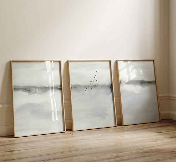 Three rectangular, light wood-framed prints featuring abstract landscape scenes in muted shades of gray and white are displayed leaning against a flat, off-white wall. They are resting on a light-colored, wood plank floor. The prints feature soft, blended watercolor-like textures with a suggestion of a horizon line and faint, silhouetted birds in the middle print. The frames are thin and simple.