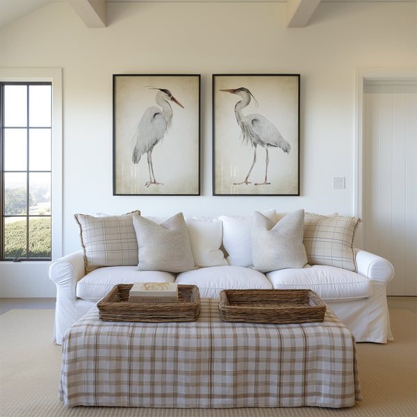 A pair of framed watercolor paintings depicting elegant great blue herons in soft neutral tones against a vintage-inspired background. The paintings are displayed on a white wall above a comfortable sofa in a living room with a view.
