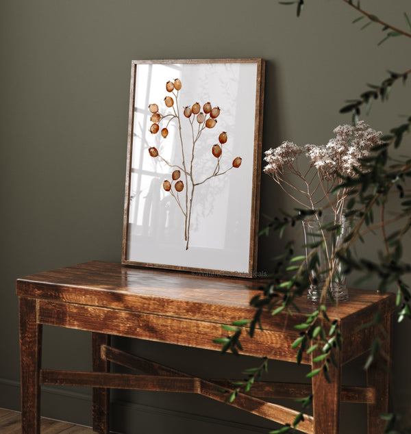 A framed botanical print featuring a delicate branch with dried rose hips in shades of brown and gold hangs on a wooden table in a cozy room with a dark green wall.