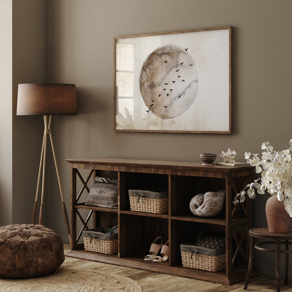 A framed watercolor painting of a moon in warm brown and beige tones with birds flying around it hangs above a wooden console table in a cozy living room with a neutral color palette.