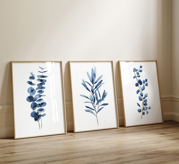 Three navy blue watercolor botanical prints of eucalyptus and leaf branches leaning on a wooden floor