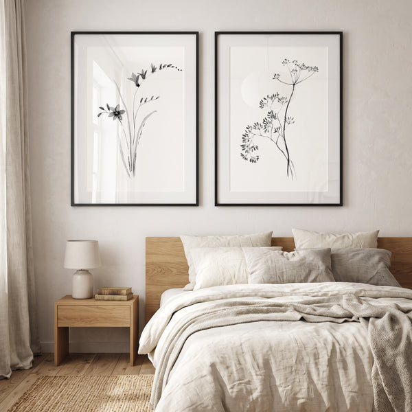 Two framed botanical art prints with black ink sketches on white backgrounds, hung side-by-side above a bed in a minimalist, neutral-toned bedroom.