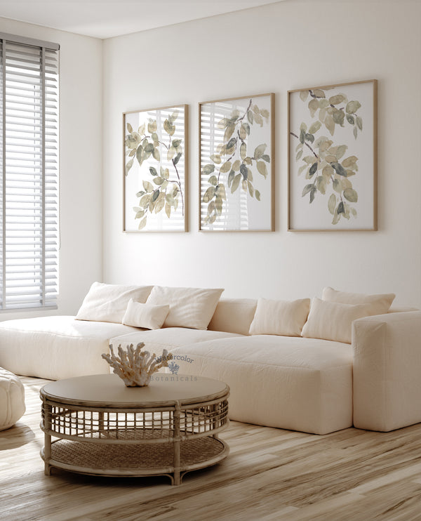 A triptych of watercolor paintings featuring delicate branches and leaves in soft shades of green and brown. The paintings are framed and displayed on a white wall above a modern sectional sofa.