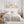 A styled bedroom scene with a bed dressed in cream and beige linens, flanked by matching nightstands and lamps, with three framed botanical prints above the headboard. A wicker basket and folded throw sit on the floor nearby.