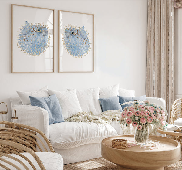 Set of 2 blue puffer fish wall art prints in wooden frames – coastal watercolour sea life artwork styled above white sofa in bright living room with rattan accents and pastel flowers
