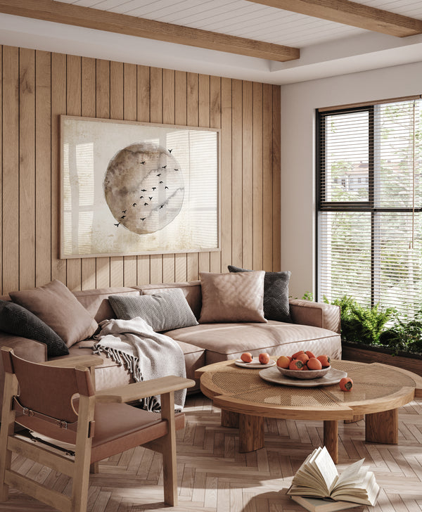 A framed watercolor painting of a moon  in warm brown and beige tones with birds flying around it hangs above a cozy sectional sofa in a living room with a neutral color palette and wooden paneling.