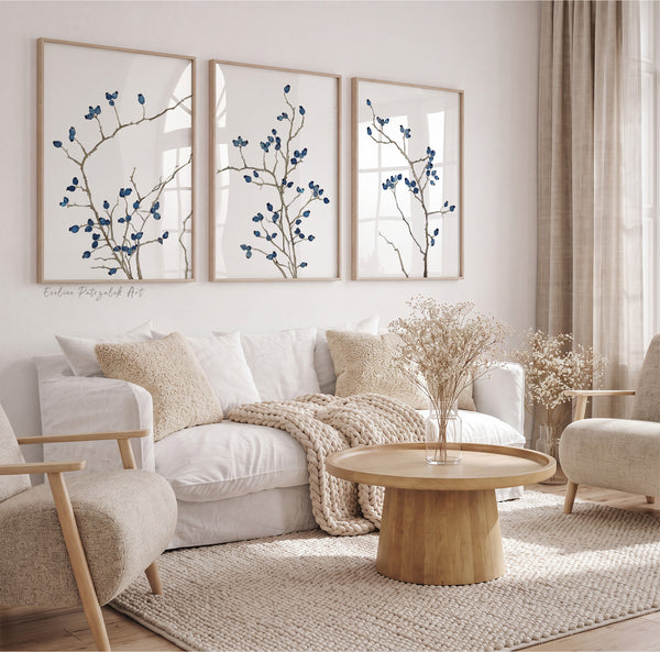 Three framed botanical wall art prints with blue branches motifs hang above a white sofa in a bright, neutral-toned living room with beige chairs, a round wood coffee table, and woven rugs.