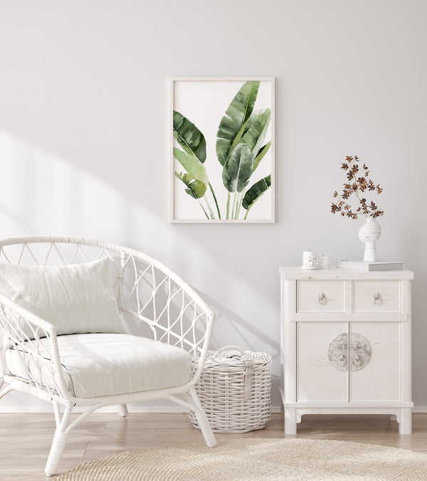 A framed watercolor painting of large green tropical banana leaves hangs on a wall above a white wicker chair in a living room.