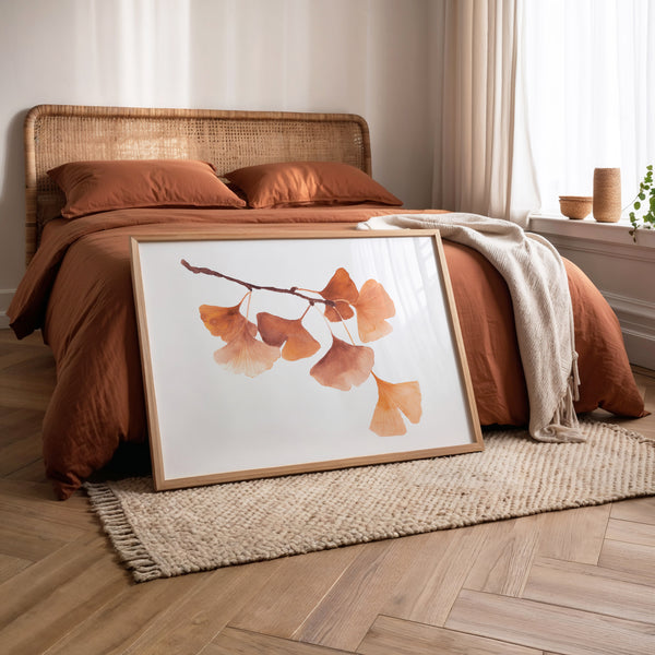 A framed watercolor art print of ginkgo leaves leaning against a bed in a warm, modern bedroom setting.